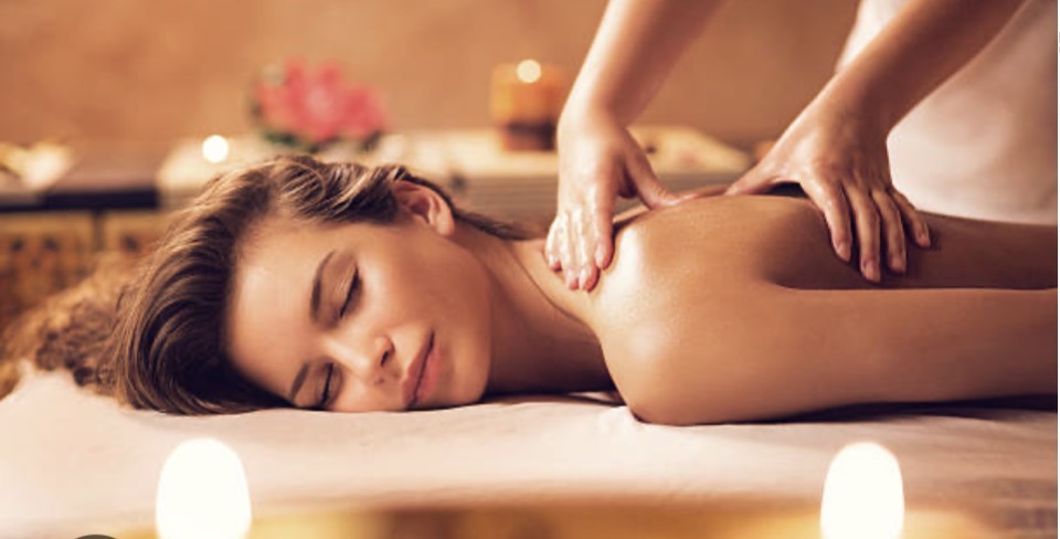 A woman lying face down on a massage table draped in white linen, eyes closed, while a therapist's hands work along her shoulder by warm candlelight.
