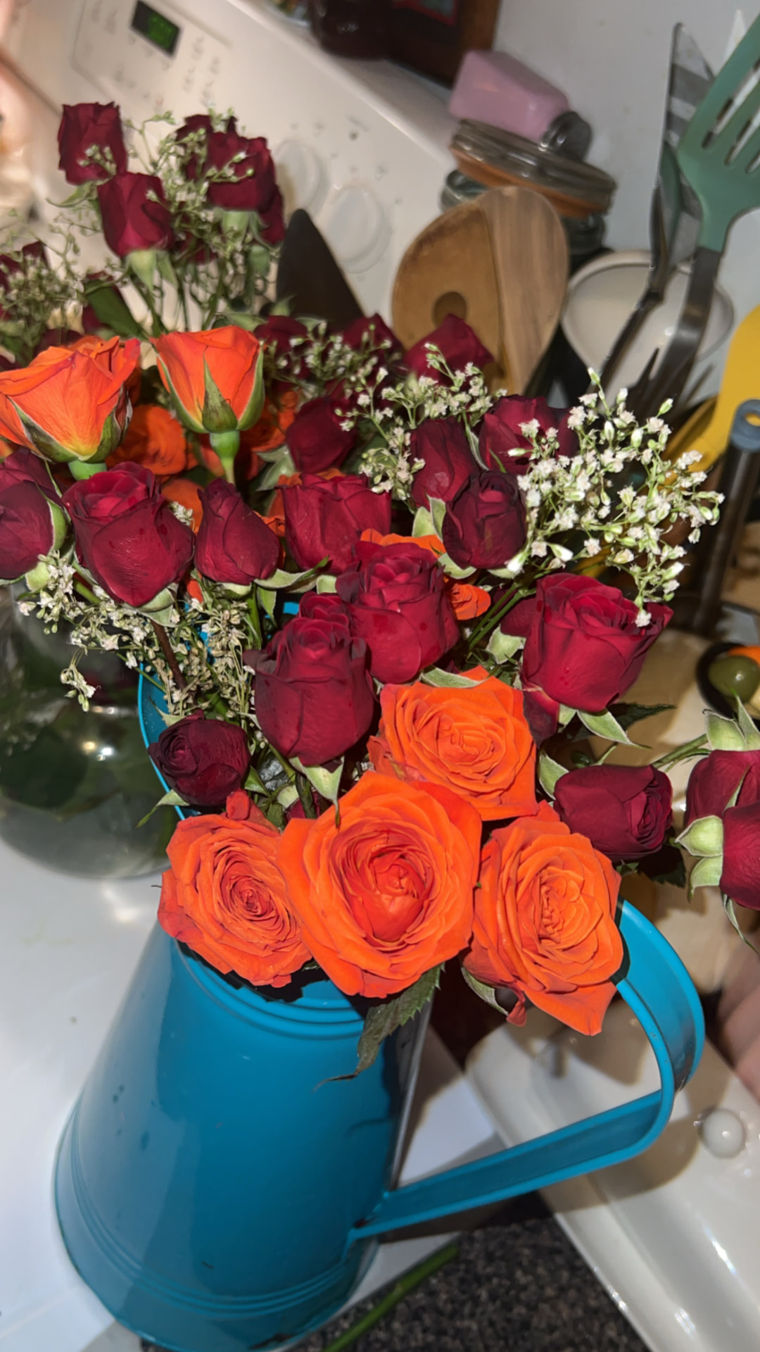 Hand-arranged bouquet of orange and deep red spray roses with baby's breath in a teal metal watering can — a Cheeky Blooms arrangement.